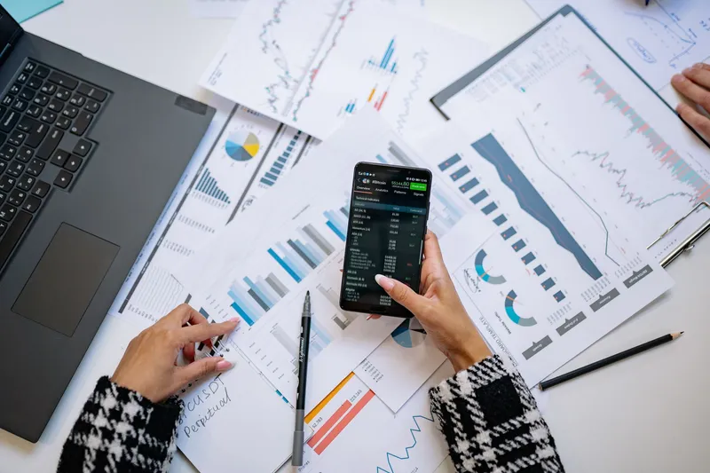Overhead view of a desk covered in printed financial charts, with a laptop and a phone showing performance figures